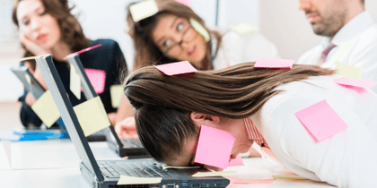 A woman lying facedown on her laptop with pink post-it notes stuck to her face, with other people in the background who have post-it notes stuck on them.