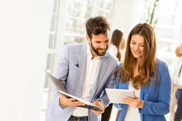 A male and female coworker comparing notes on a tablet and a clipboard smiling.
