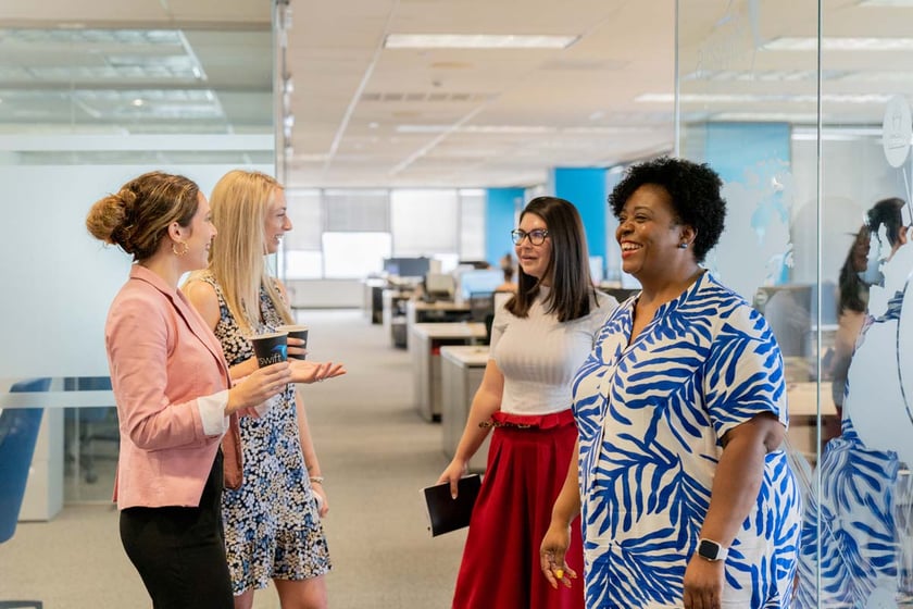 Four women chatting in a modern office: one with a coffee cup, another in floral, one with glasses and a portfolio, and one in a striped blouse.