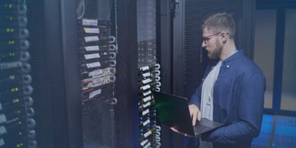 A data center technician stands inside a server room, working on a laptop while inspecting network racks and hardware components, a typical task in data center jobs and operations careers.