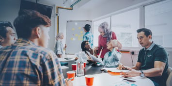 A group of multicultural colleagues actively engaged in a team meeting. Two employees stand by a whiteboard filled with sticky notes, while others are seated around a table discussing documents and data. The scene captures collaboration and brainstorming during a training session.