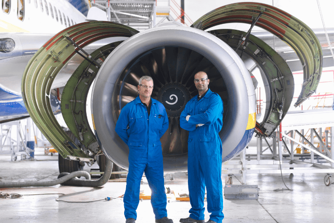 Two aerospace engineers in blue work overalls standing in front of a large commercial jet engine
